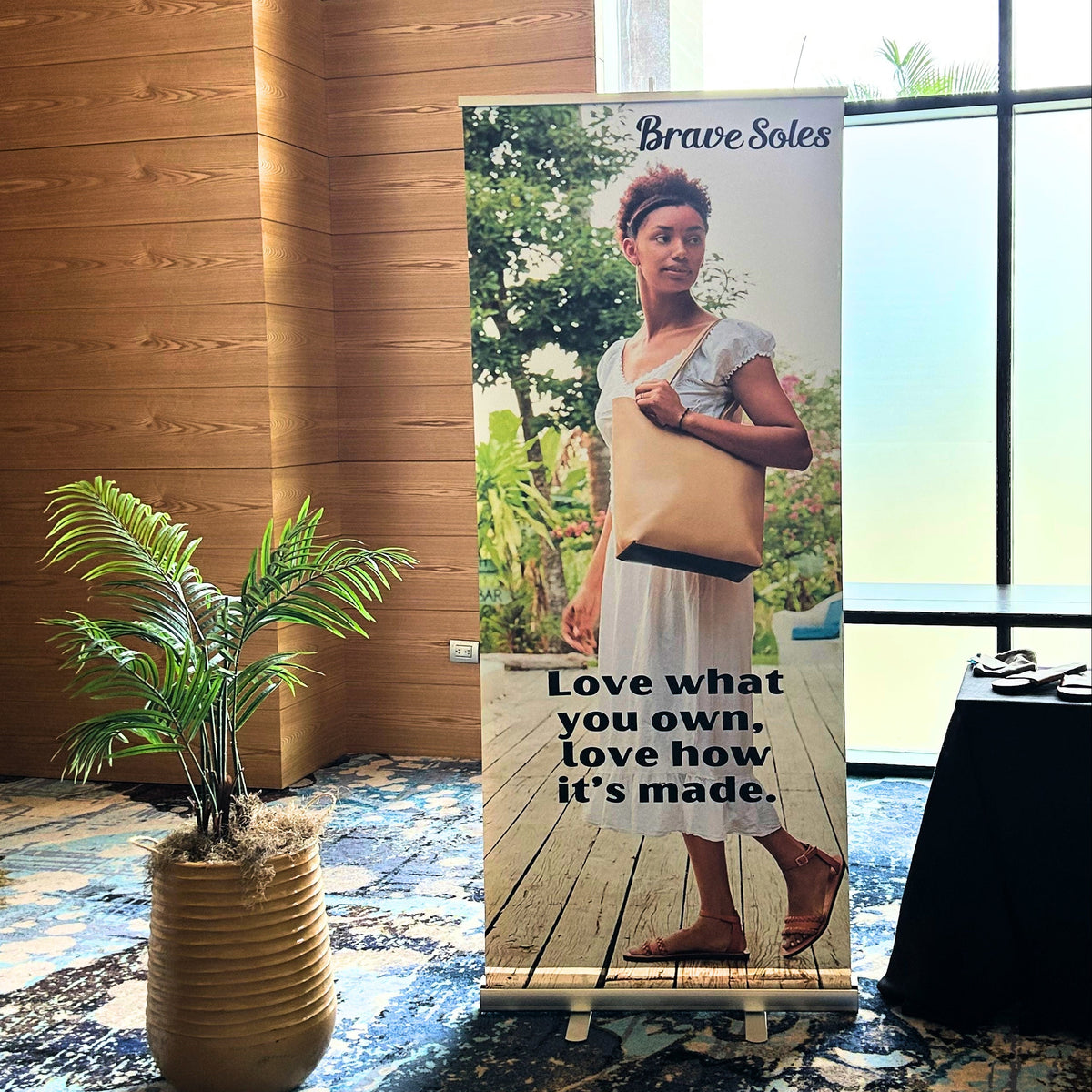 A vertical Brave Soles branded roll-up banner stand on a small easel in an indoor event space. The banner features a model wearing a beige dress and brown sandals, with the text 'Brave Soles' at the top and 'Love what you own, love how it's made.