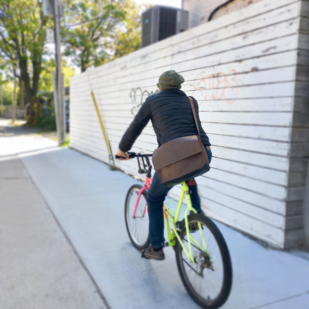 Person riding a bicycle with a Miina Goods Gabriel messenger bag on a sidewalk next to a wall with graffiti.