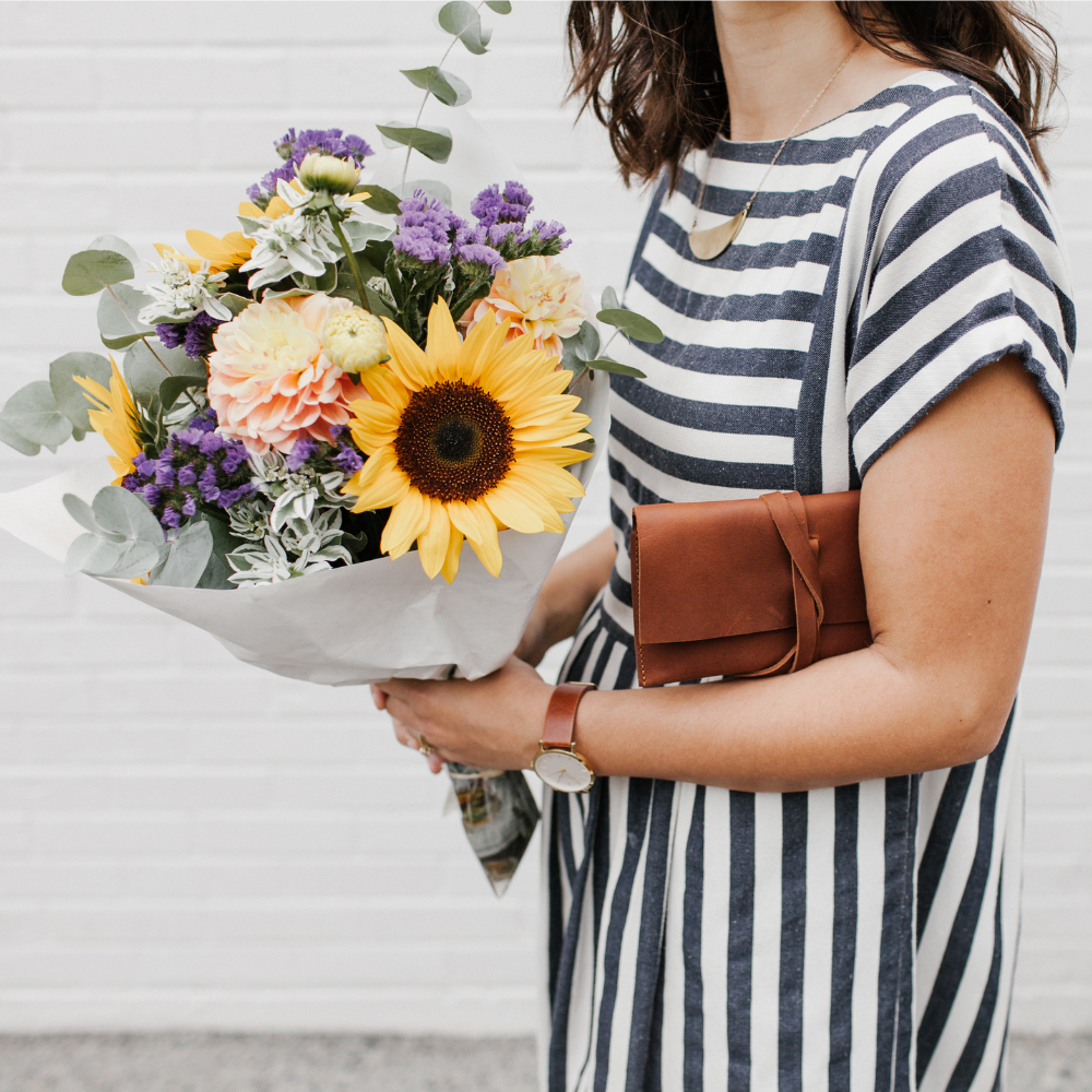A woman wearing a navy and white striped t-shirt dress holds a large bouquet of colorful flowers, including a prominent yellow sunflower, next to the Miina Sandra leather clutch wallet in caramel. 