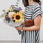 A woman wearing a navy and white striped t-shirt dress holds a large bouquet of colorful flowers, including a prominent yellow sunflower, next to the Miina Sandra leather clutch wallet in caramel. 